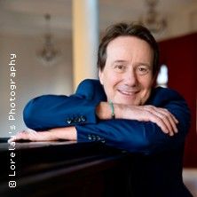 Ein lächelnder Mann in einem blauen Anzug lehnt sich auf ein Klavier.