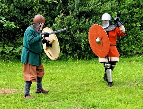 Two fighters in historical armor are practicing a battle on a meadow. One is carrying a shield and a sword, while the other is wearing armored armor and has a large shield.