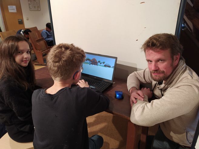 Ein Mann und zwei Kinder sitzen an einem Tisch mit einem Laptop, auf dem ein digitales Modell angezeigt wird.