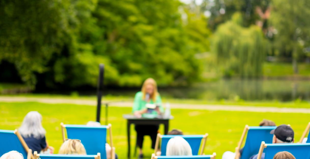 Menschen sitzen in blauen Liegestühlen in einem Park und hören einer Person an einem Tisch zu.