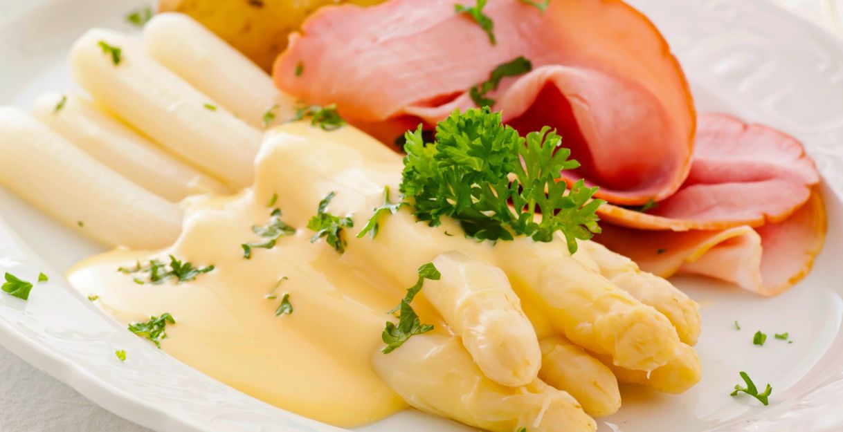 Ein Teller mit weißem Spargel, Schinken, Kartoffeln und Sauce Hollandaise, garniert mit Petersilie.