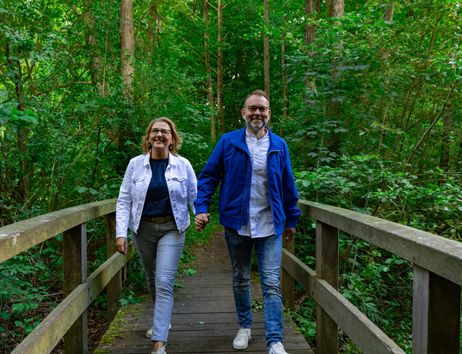 A couple is walking hand in hand across a wooden bridge in a green forest. The path is surrounded by trees and plants.