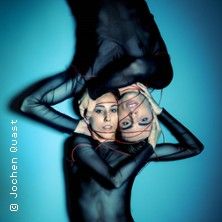 Zwei Tänzerinnen in schwarzen Outfits posieren kunstvoll auf blauem Hintergrund.