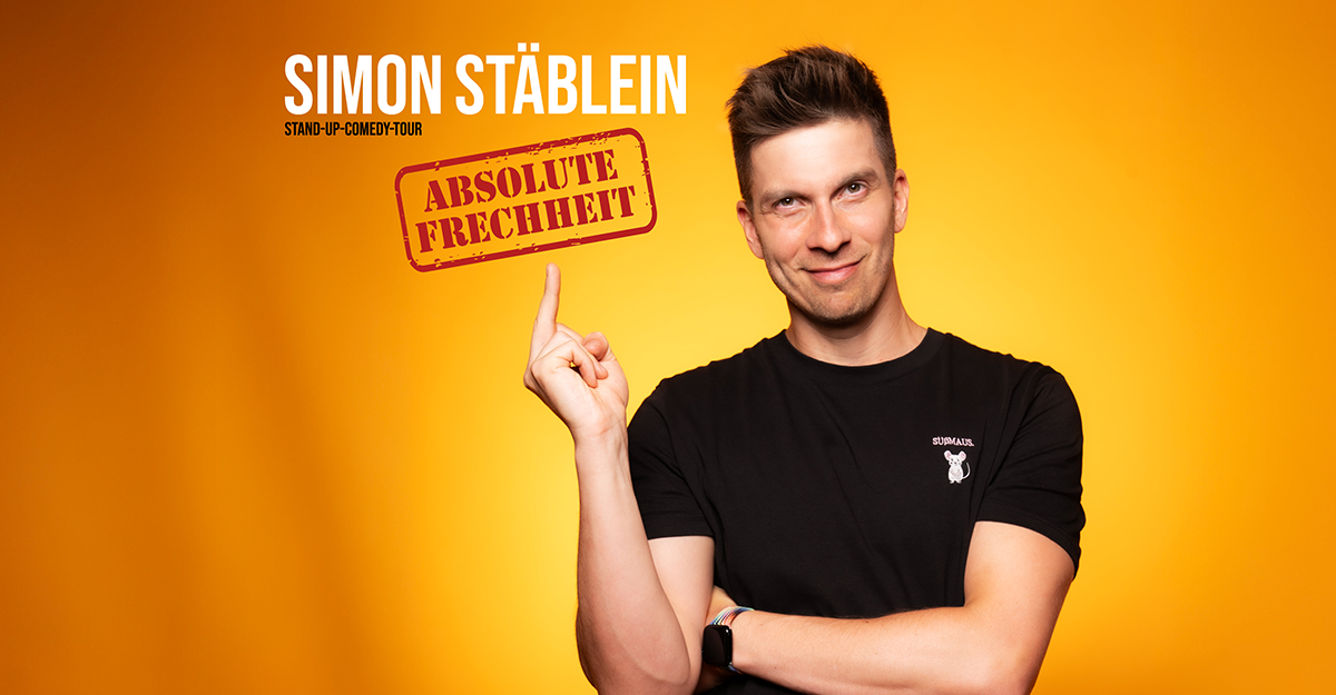 Ein Mann in schwarzem T-Shirt zeigt mit dem Finger auf den Schriftzug 'Absolute Frechheit' vor einem orangefarbenen Hintergrund.