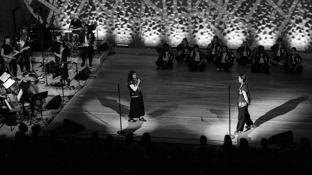 Schwarz-weiß-Foto einer Bühnenperformance mit zwei Personen im Vordergrund und einer Band im Hintergrund.