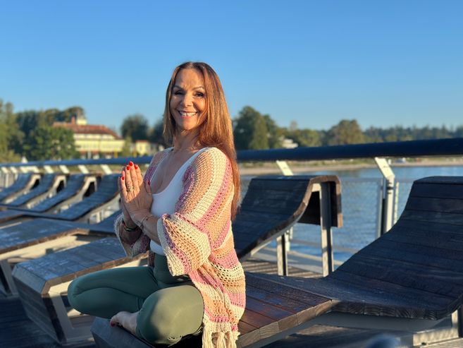 Eine Frau meditiert auf einer Holzliege an einem sonnigen Tag auf einer Seebrücke.