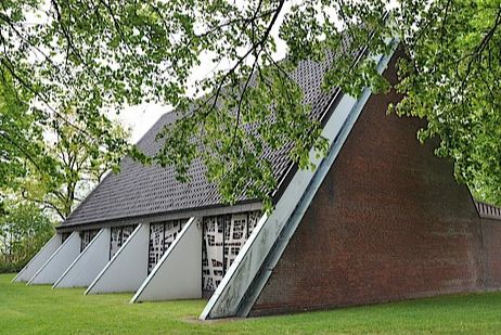 Moderne Kirche mit schrägen Wänden und großen Fenstern, umgeben von Bäumen.