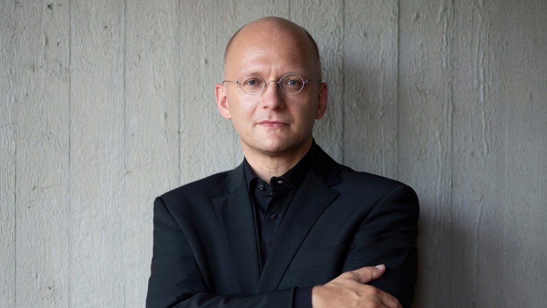 Ein Mann in schwarzem Anzug steht vor einer grauen Wand.