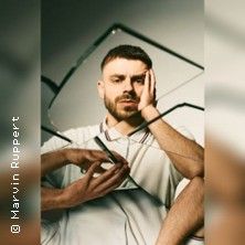 Ein Mann mit Bart hält ein zerbrochenes Spiegelstück vor sein Gesicht. Er trägt ein weißes Hemd und schaut nachdenklich.