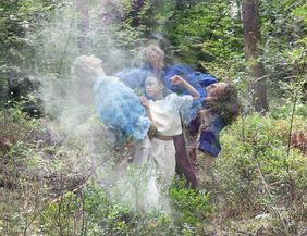 Mehrere Personen in blauer Kleidung in einem Wald, umgeben von Nebel.