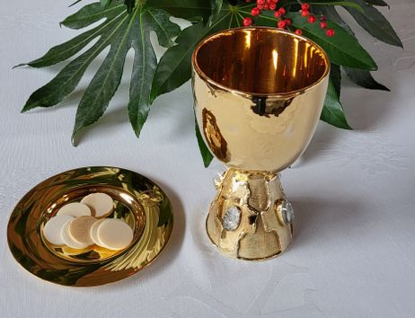 Goldener Kelch und Patene mit Hostien auf einem weißen Altartuch, umgeben von grünen Blättern und roten Beeren.