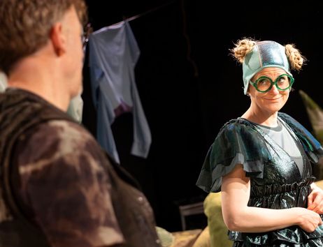 Ein Mann ist von hinten zu sehen. Er guckt eine Frau (Antje Otterson) an, die ein schillerndes Kleid und eine schillernde Badehaube trägt. Sie lächelt ihn verschämt an.