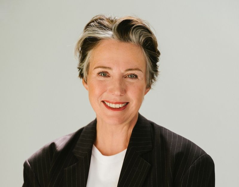 Porträt einer lächelnden Frau mit kurzem Haar in einem dunklen Anzug vor hellem Hintergrund.