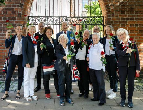 Eine Gruppe von Menschen hält rote Rosen vor einem Backsteinbogen.