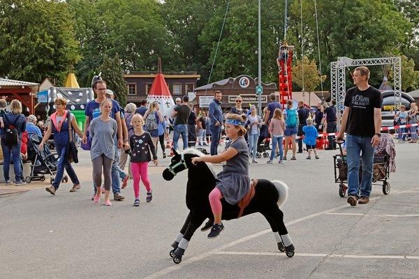 A cheerful folk festival with many people and stalls. A child rides on a toy horse and attracts the attention of passersby.
