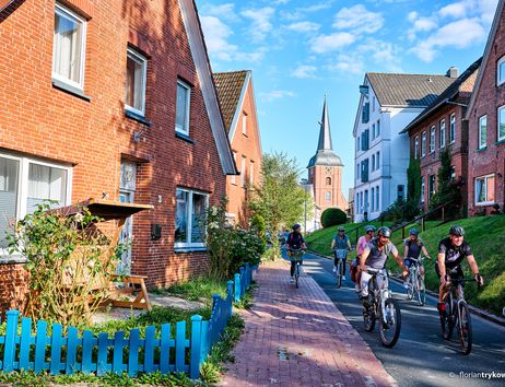 Gruppe von Radfahrern auf einer Straße mit Backsteinhäusern und Kirchturm im Hintergrund.