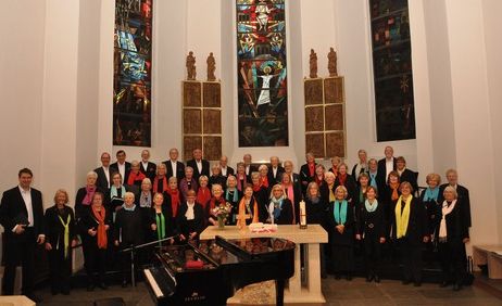 Ein Chor posiert in einer Kirche vor bunten Glasfenstern und einem Klavier.