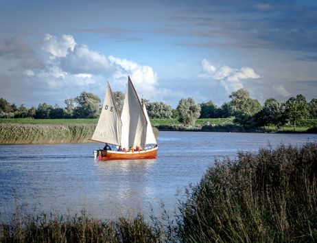 Ein Segelboot mit weißen Segeln fährt auf einem Fluss, umgeben von grüner Landschaft und blauem Himmel.