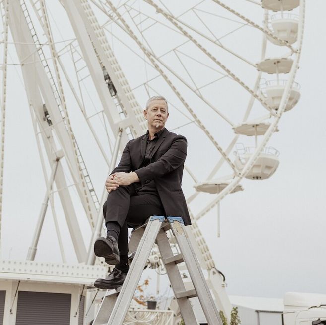 Eine Person in schwarzem Anzug sitzt auf einer Leiter vor einem Riesenrad.