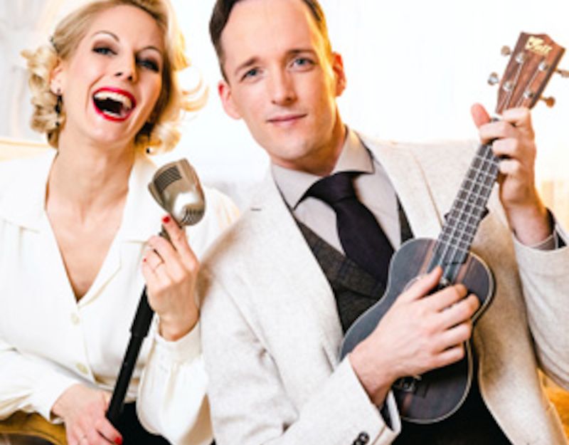 Ein Mann und eine Frau in eleganter Kleidung, die Frau hält ein Mikrofon, der Mann eine Ukulele.