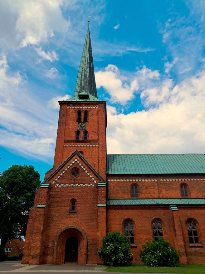 A tall brick church with a pointed tower and a green roof. The sky is blue with some clouds.