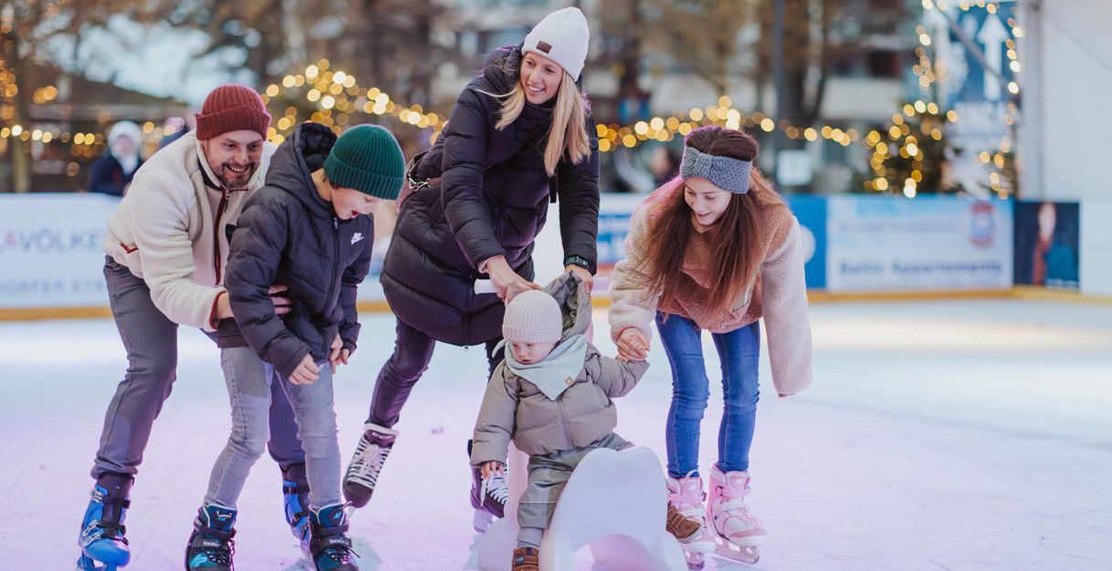 Eine Familie beim Schlittschuhlaufen auf einer Eisbahn, umgeben von Lichterketten.