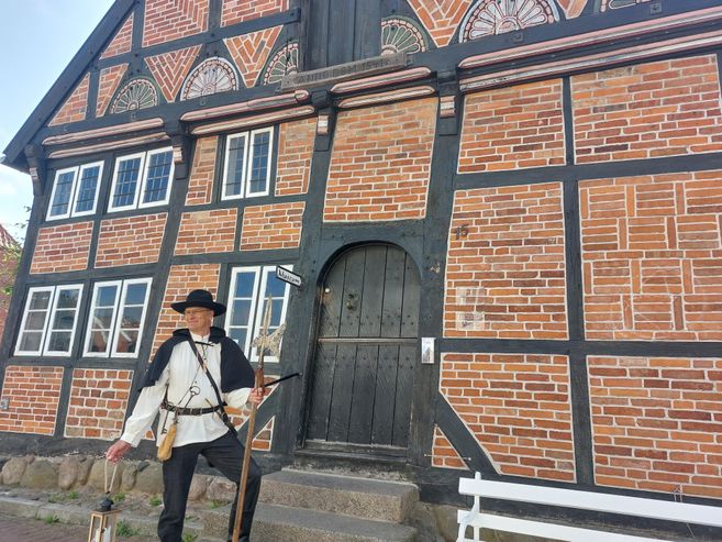 A historical timber-framed house with reddish-brown bricks and black beams. In front of the house stands a person in traditional clothing.