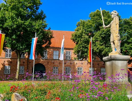 Statue vor einem historischen Gebäude mit Flaggen und Blumenbeet in Bad Bramstedt.