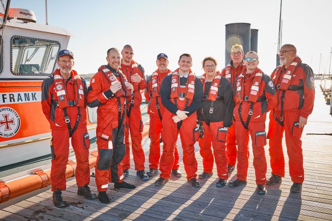 Eine Gruppe von Menschen in roten Rettungsanzügen steht auf einem Steg vor einem Rettungsboot.
