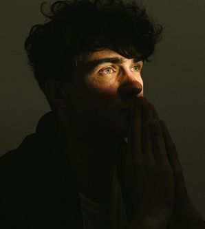 Ein Mann im Halbschatten mit nachdenklichem Blick, Hände vor dem Gesicht gefaltet.