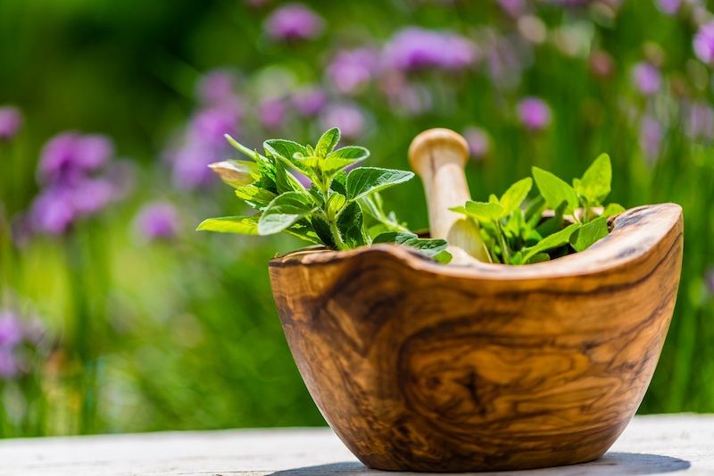 Holzmörser mit frischen Kräutern im Freien vor unscharfem Hintergrund mit lila Blumen.
