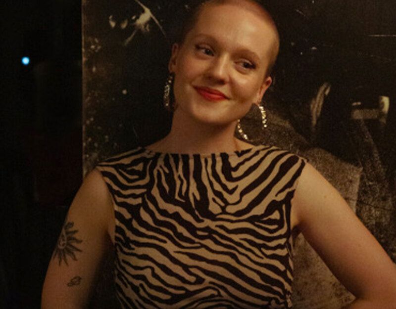 Person mit kurzem Haar und Zebra-Muster-Kleid lächelt vor dunklem Hintergrund.