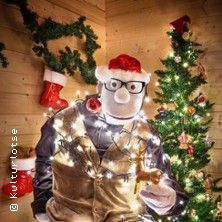 Puppe mit Weihnachtsmütze und Lichterkette vor Weihnachtsbaum.