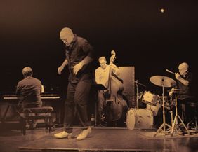 Ein Musikertrio auf der Bühne mit einem Stepptänzer im Vordergrund, einem Pianisten, einem Kontrabassisten und einem Schlagzeuger im Hintergrund.