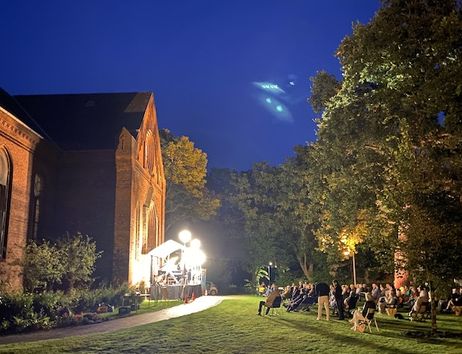 Open-Air-Konzert auf einer Wiese neben einem Backsteingebäude bei Nacht.