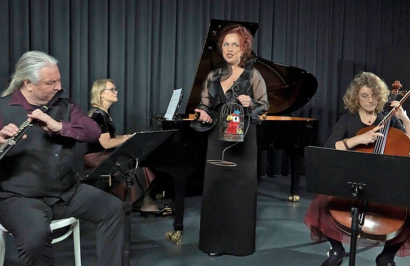 Musiker spielen klassische Musik mit Flöte, Klavier und Cello. Eine Frau hält einen Vogelkäfig.