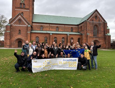 A group of people stands in front of a church holding up a banner. The banner displays information about an event in the city center.
