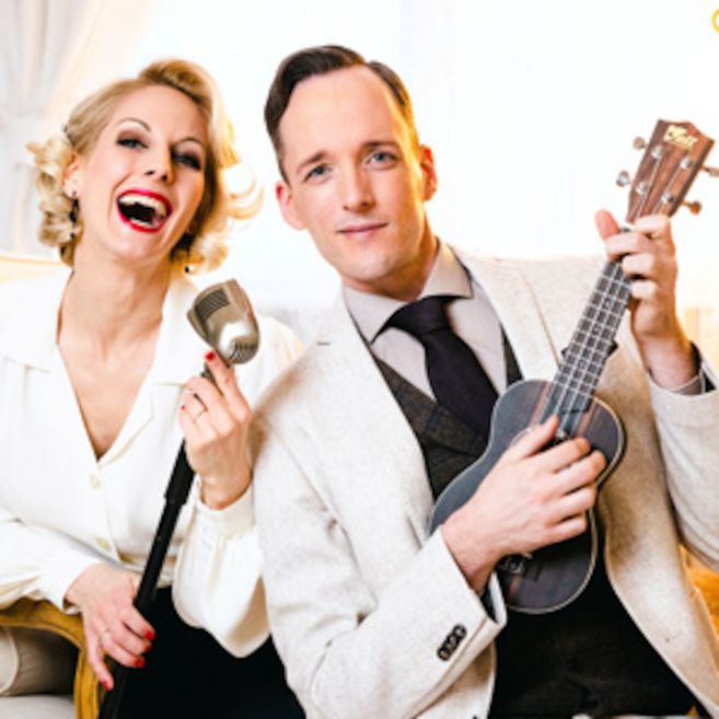 Ein Mann und eine Frau in eleganter Kleidung, die Frau hält ein Mikrofon, der Mann eine Ukulele.