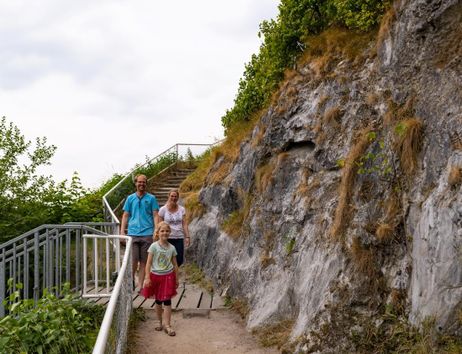 A family is walking along a path beside a steep rock face. In the background, there are stairs and a lot of green foliage visible.