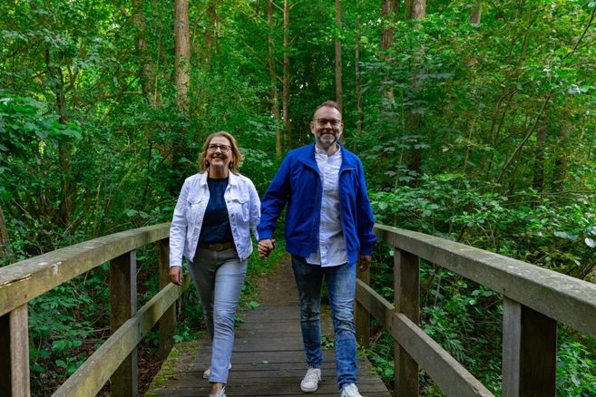 A couple is walking hand in hand across a wooden bridge in a green forest. The path is surrounded by trees and plants.