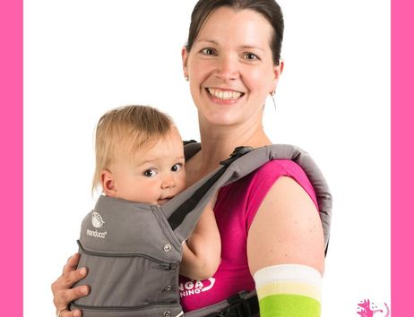 Frau mit Baby in einer Trage, lächelnd, vor weißem Hintergrund mit pinkem Rahmen und Kanga Training Logo.