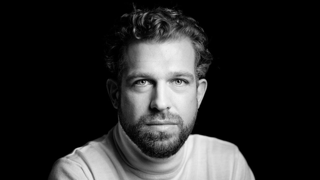 Schwarz-weiß Porträt eines Mannes mit Bart und lockigem Haar, der direkt in die Kamera schaut.