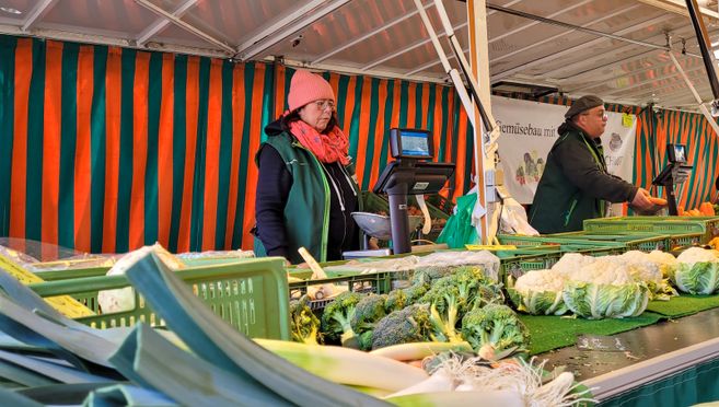 Zwei Verkäufer an einem Gemüsestand auf einem Wochenmarkt mit Brokkoli und Blumenkohl im Vordergrund.