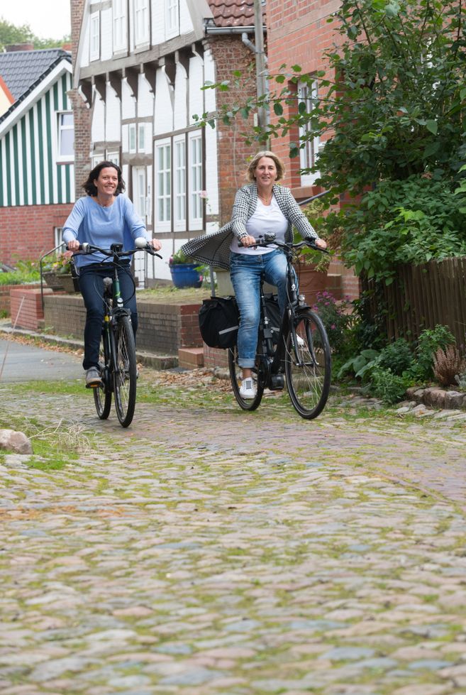 Zwei Frauen fahren auf Fahrrädern eine gepflasterte Straße entlang, umgeben von traditionellen Häusern.
