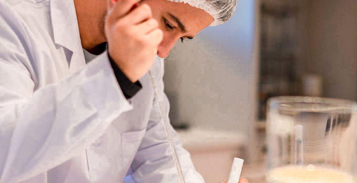 Person in weißem Laborkittel und Haarnetz arbeitet mit Laborgeräten und Flüssigkeiten.