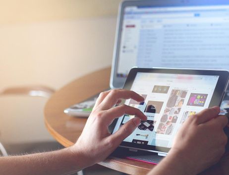 Person nutzt ein Tablet mit einem Laptop im Hintergrund auf einem Tisch.