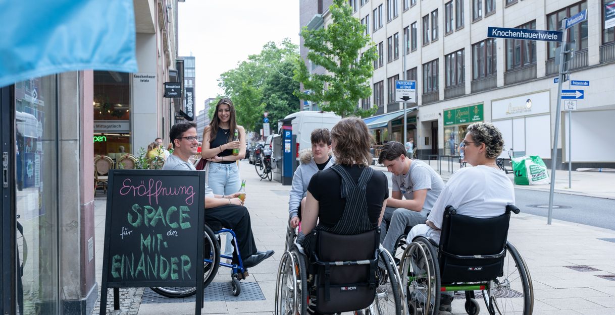 Menschen, die im Rollstuhl sitzen und eine Person, die steht, im Gespräch miteinander