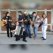 Eine sechsköpfige Band spielt Instrumente vor einer Backsteinwand.