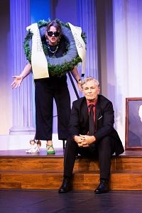 Zwei Personen auf einer Bühne, eine sitzt, die andere steht mit einem Kranz um den Hals.