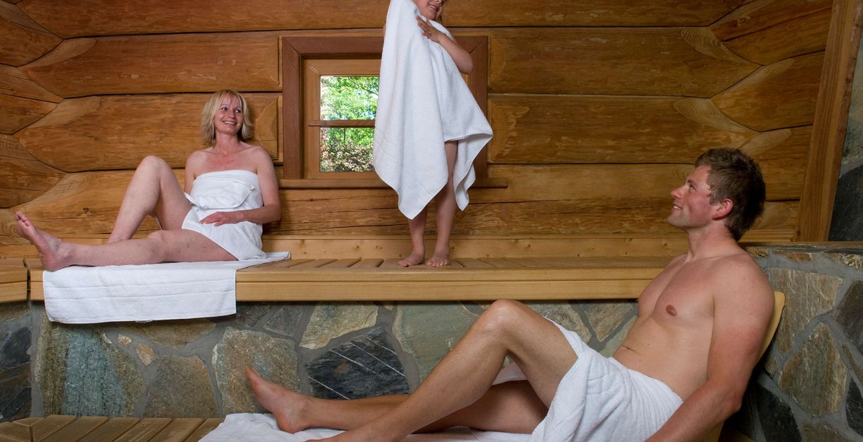 Eine Familie entspannt in einer Holzsauna. Ein Kind steht in der Mitte mit einem Handtuch, während die Eltern auf den Bänken sitzen und liegen.